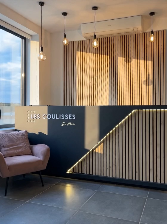 Comptoir d'accueil avec le logo Les Coulisses de Marie, bois clair et suspensions lumineuses.